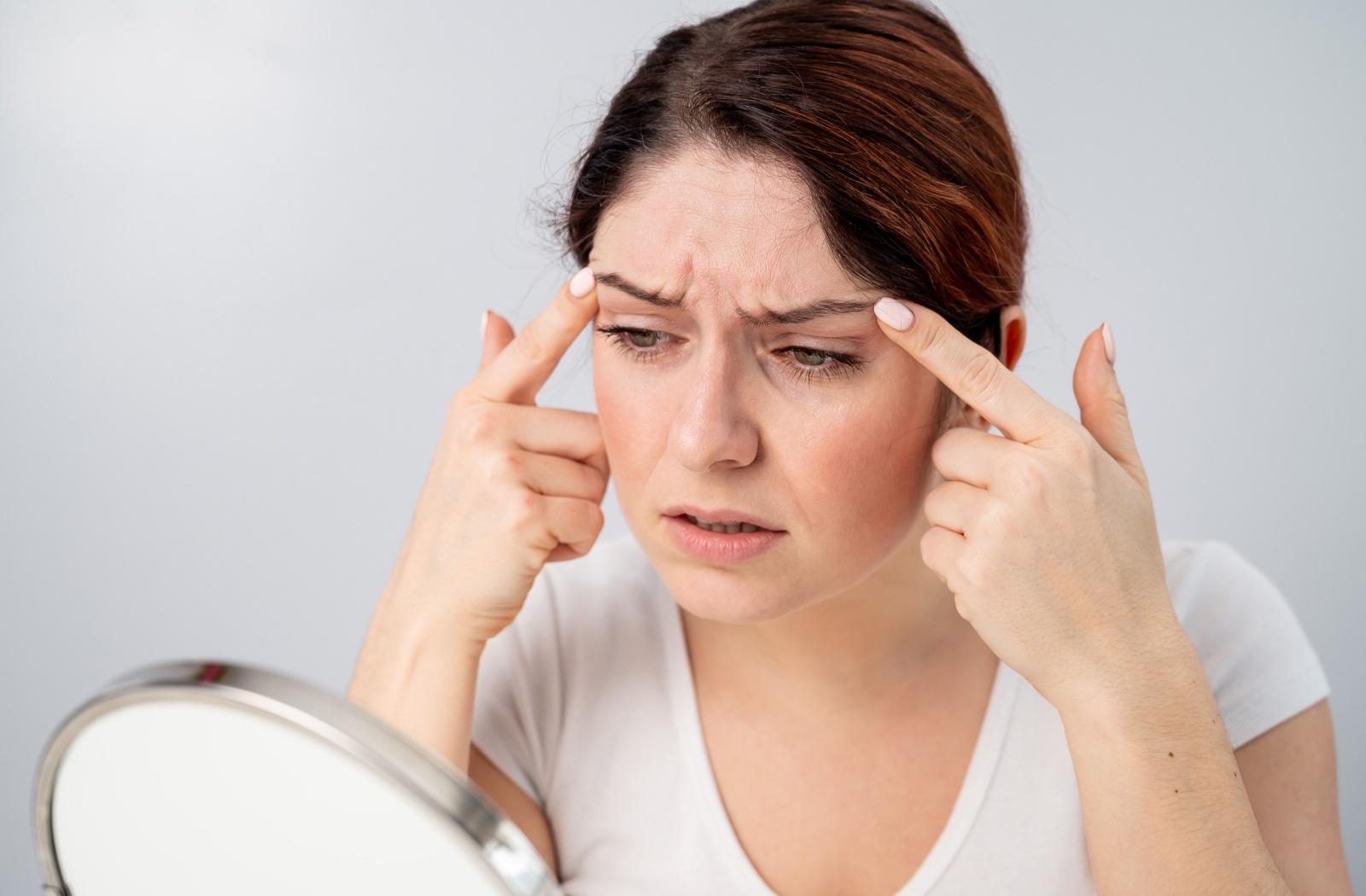 A frustrated woman touching her forehead and looking at her frown lines in the mirror.