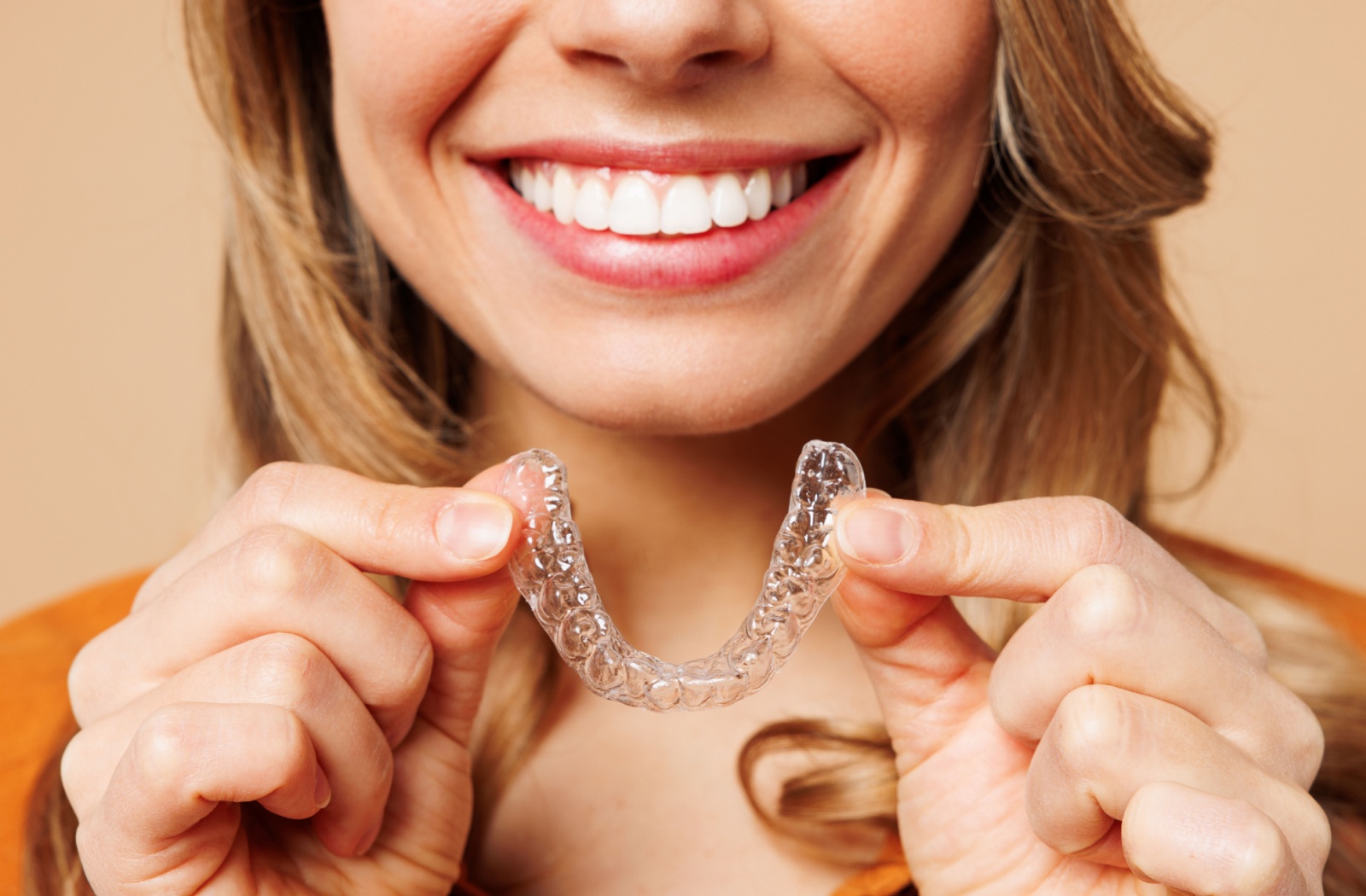 Close-up of the lower half of a person's face smiling while holding up an Invisalign aligner.