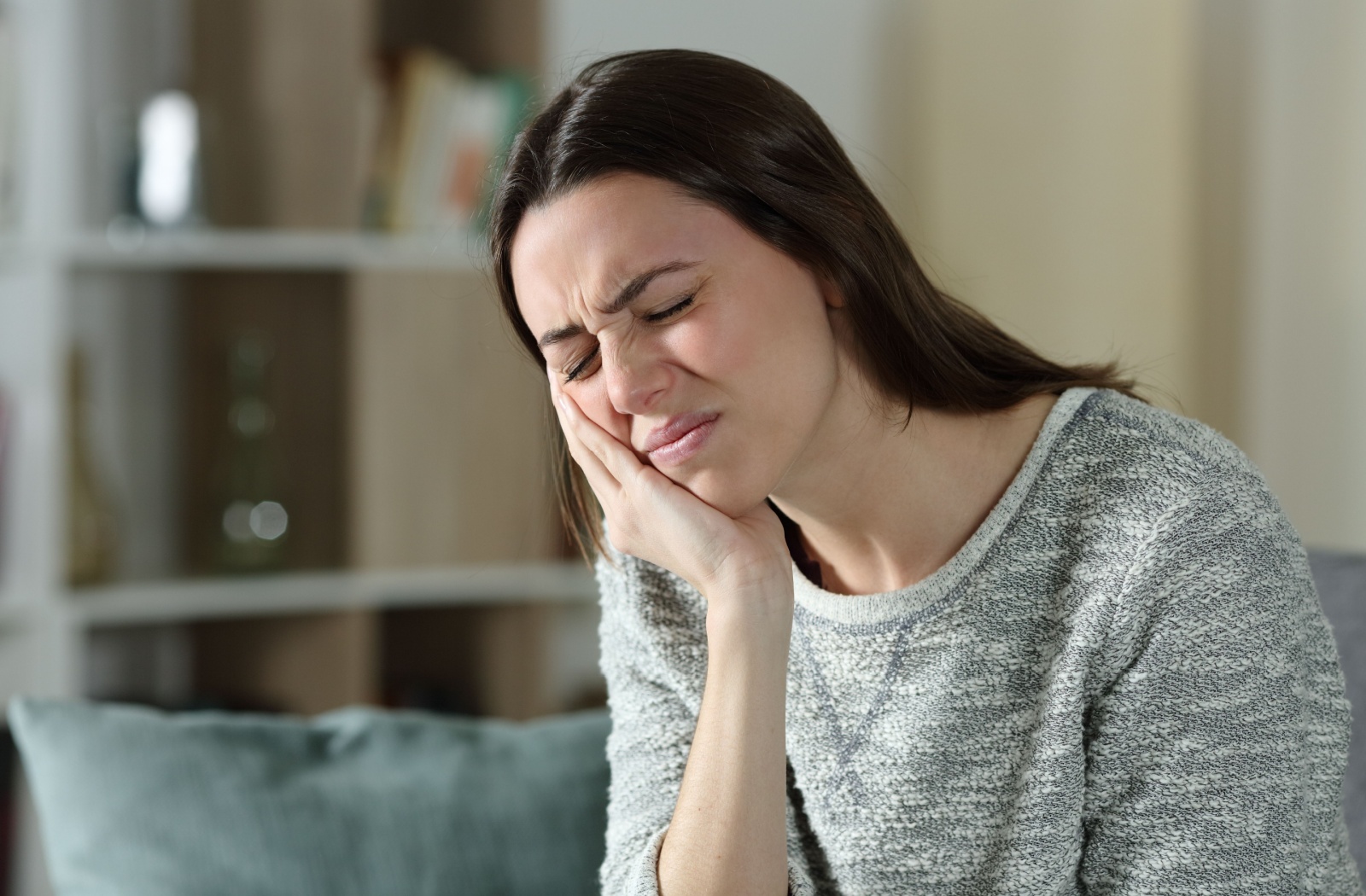 A young adult sitting on her couch rubbing her jaw and squinting in discomfort due to her TMJ disorder.