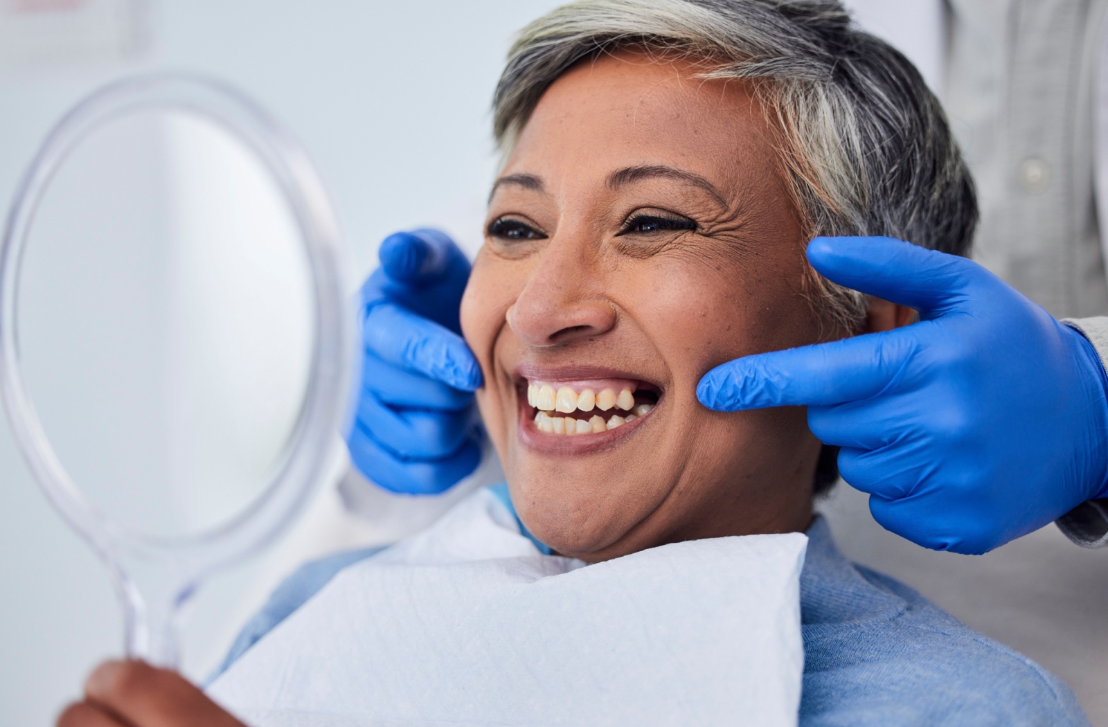 An older patient smiling in a mirror after a dental procedure while a gloved dentist points at her new dental bridge.