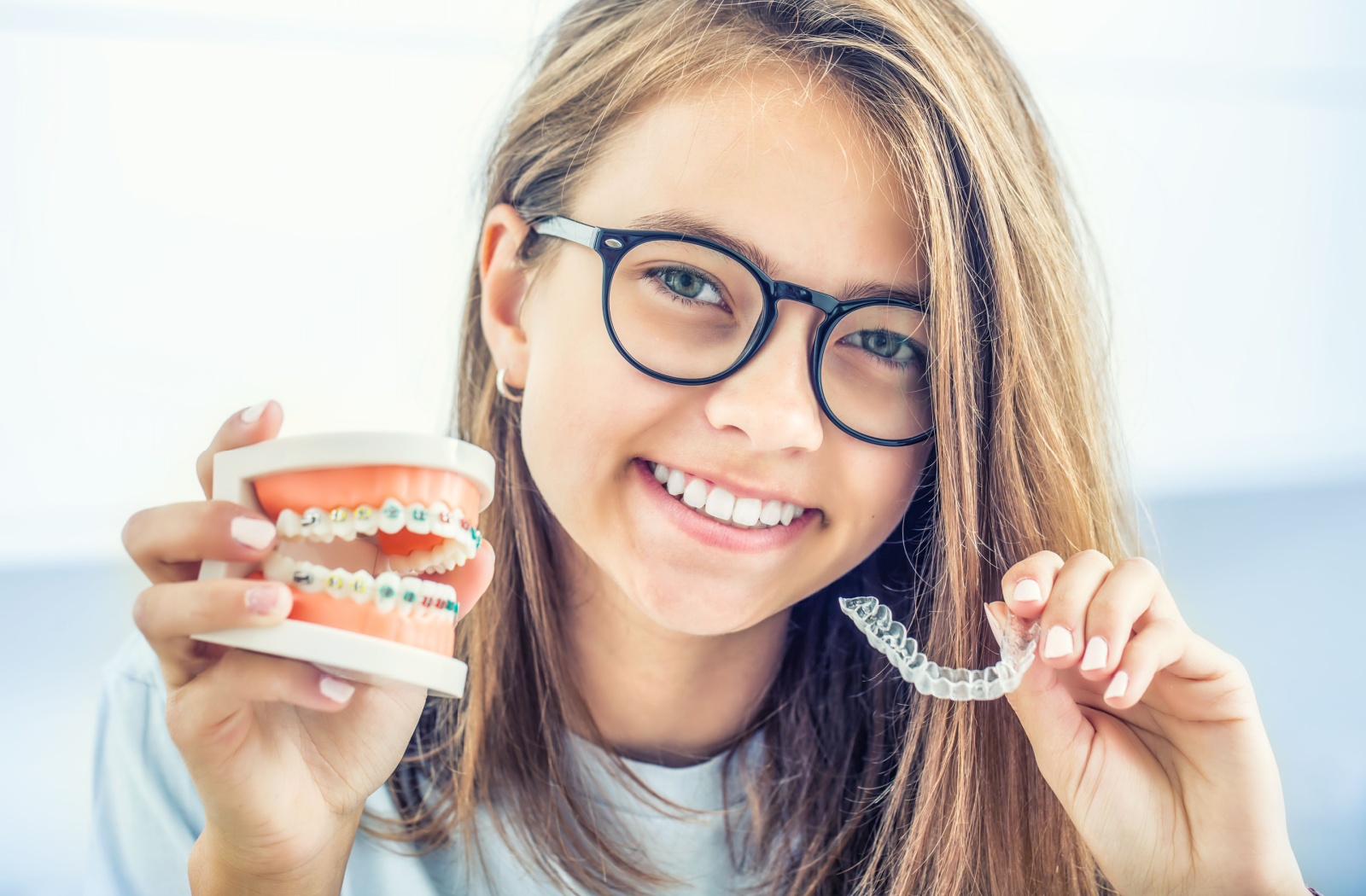 A close-up of a smiling young adult holding Invisalign in one hand and a silicone model of teeth with braces in the other.