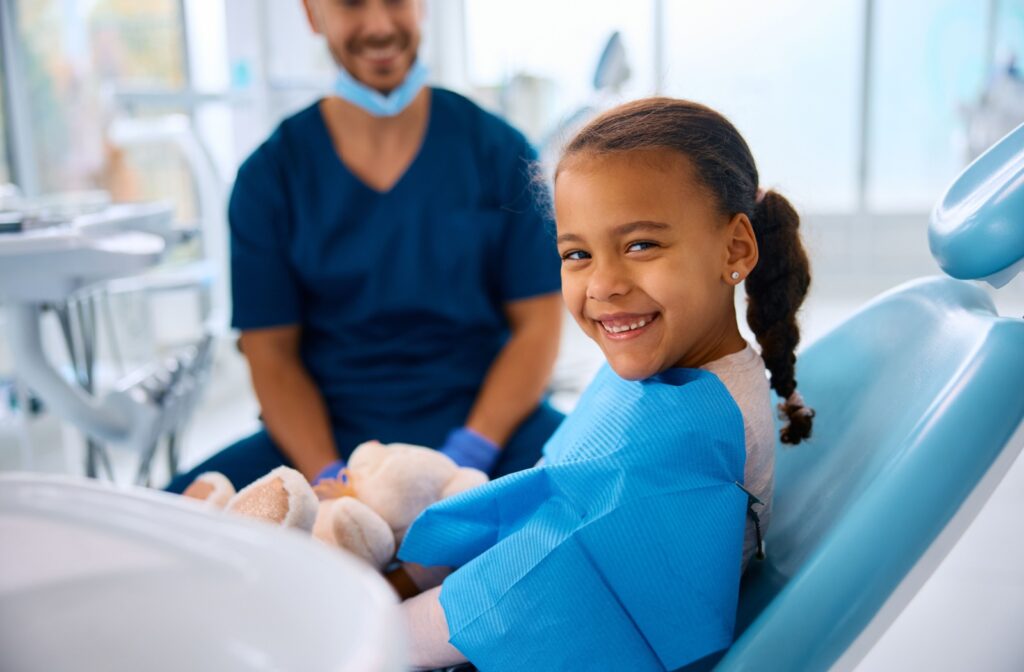 Small child sits in a dental exam chair and turns to face the camera. They have a blue dental bib around their neck, and a dentist in blue scrubs sits beside them. They both are smiling.