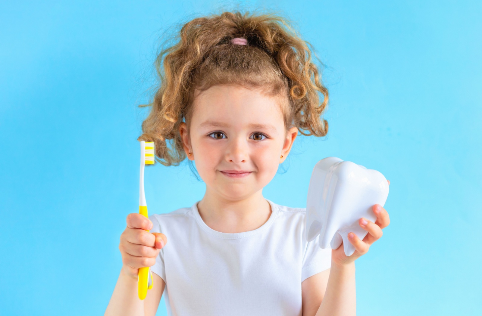 A small child wearing a white t-shirt stands in front of a blue wall, holding a toothbrush in their left hand and a model tooth in their right hand while smiling